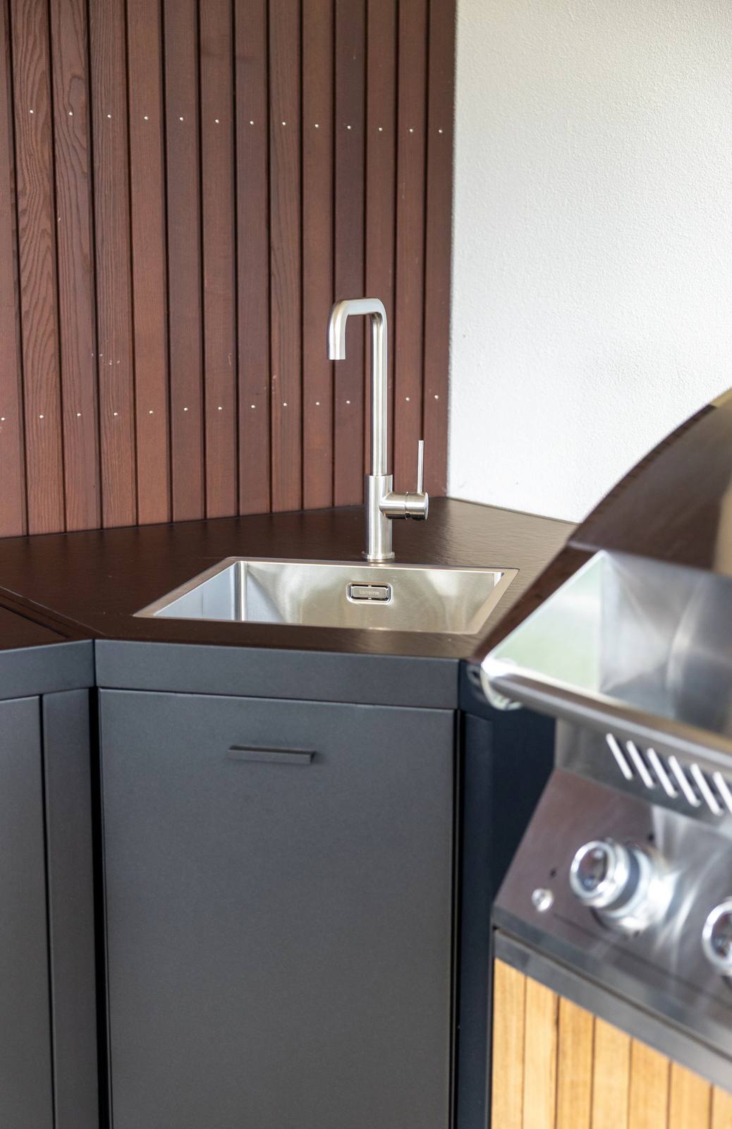 Moderne buitenkeuken met spoelbak, RVS kraan en donkere kastjes tegen een houten lattenwand, naast een barbecue